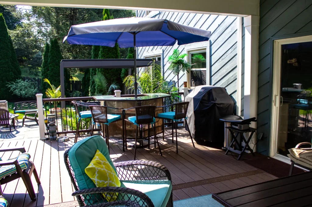 Photo of a beach-style deck featuring a glass table with an umbrella and some chairs, as well as a covered grill